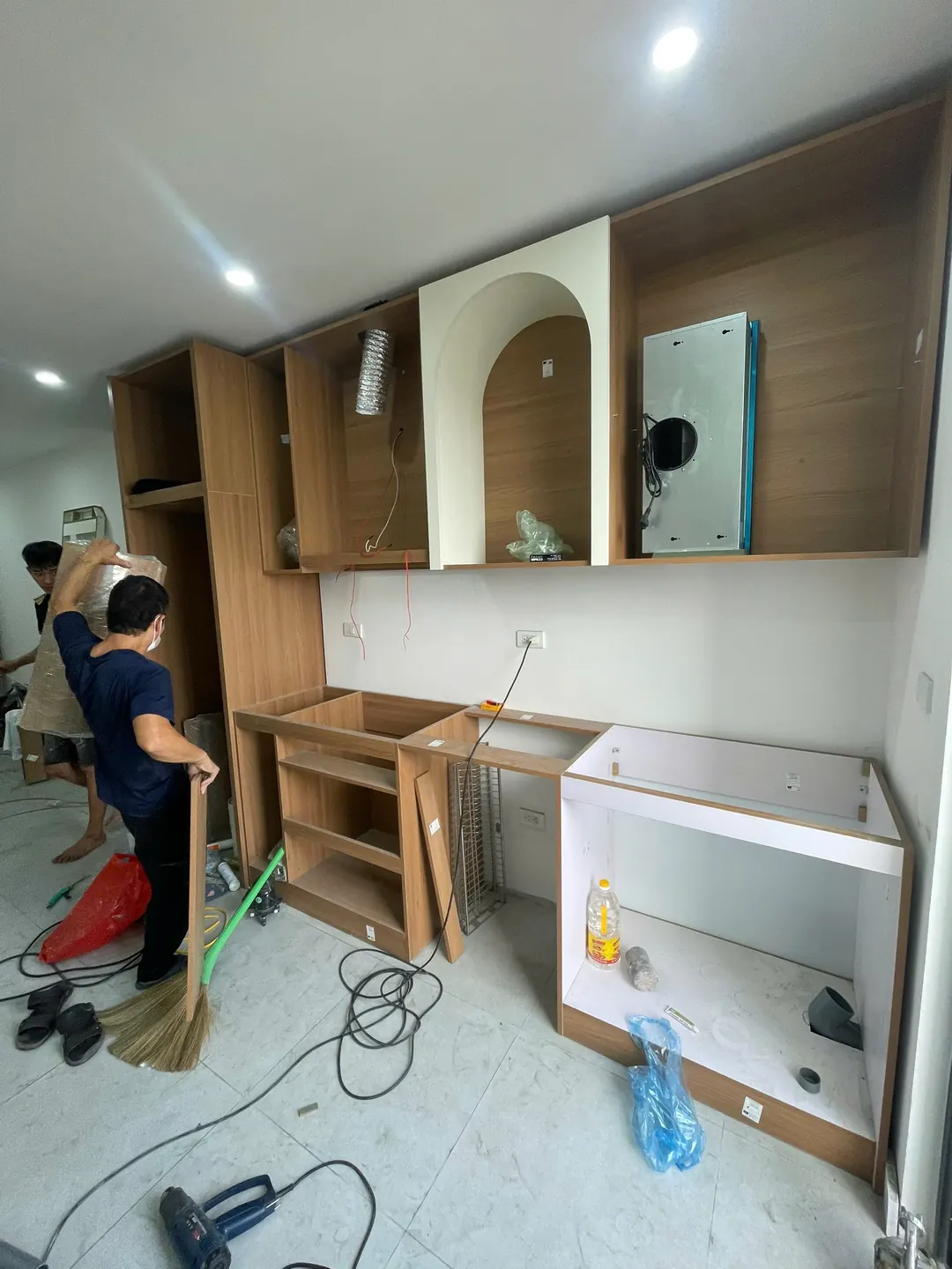 Tủ bếp trên và dưới bằng gỗ vân sáng đang lắp, tủ trên có hốc vòm trắng, thợ dọn sàn