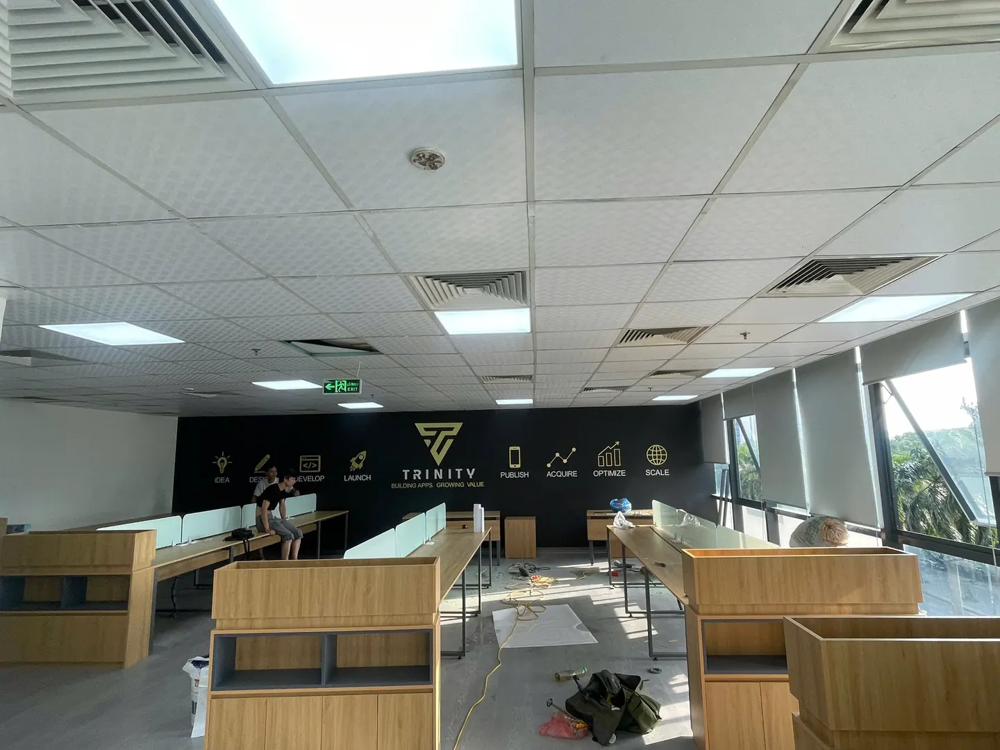 Toàn cảnh khu làm việc chung: các dãy bàn dài, vách ngăn, tủ gỗ thấp và tường logo